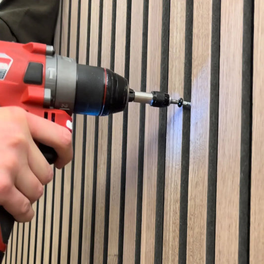 Close-up of a person using a red power drill to screw into a wooden wall with dark gray vertical panels.